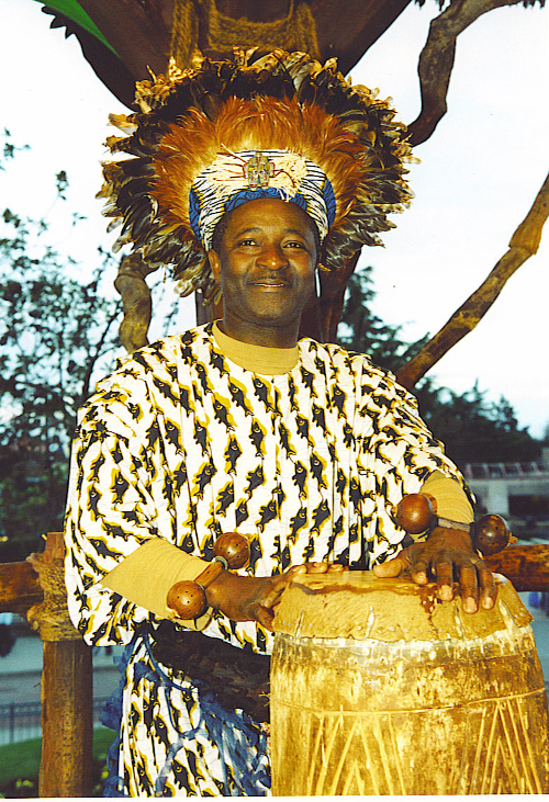 Maître Nono jouant du tam tam en costume traditionnel avec des plumes sur la tête